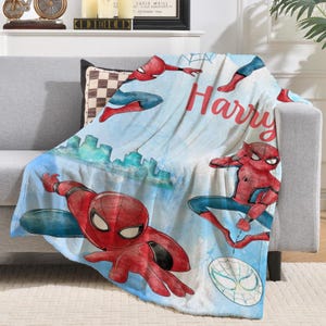 K&ouml;nnte beinhalten: Eine blau-wei&szlig;e Decke mit Illustrationen von Spider-Man in verschiedenen Posen. Die Decke hat ein Stadtbilddesign und den Namen "Harry" in roter Schrift.
