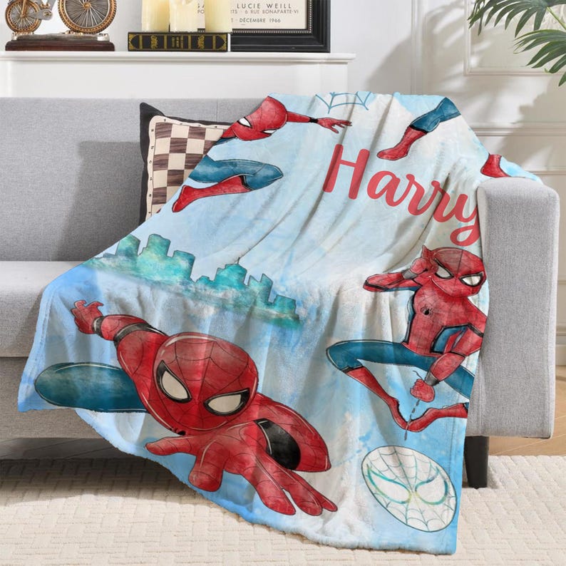 K&ouml;nnte beinhalten: Eine blau-wei&szlig;e Decke mit Illustrationen von Spider-Man in verschiedenen Posen. Die Decke hat ein Stadtbilddesign und den Namen "Harry" in roter Schrift.