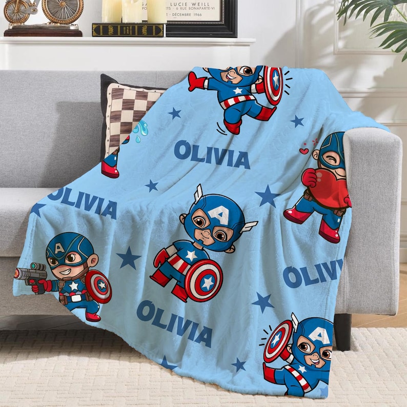 Pode incluir: Um cobertor azul claro com ilustra&ccedil;&otilde;es de desenhos animados do Capit&atilde;o Am&eacute;rica e o nome "OLIVIA" impresso em branco. O cobertor, com um padr&atilde;o de estrelas, est&aacute; sobre um sof&aacute; cinza.