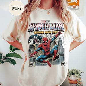 K&ouml;nnte beinhalten: Elfenbeinfarbenes T-Shirt mit einem Marvel Studios Spider-Man-Grafikdesign. Das Design zeigt Spider-Man, Venom, Stadtlandschaften und den Text "BRAND NEW DAY". Das Daily Bugle-Logo und ein Spinnensymbol sind ebenfalls zu sehen.