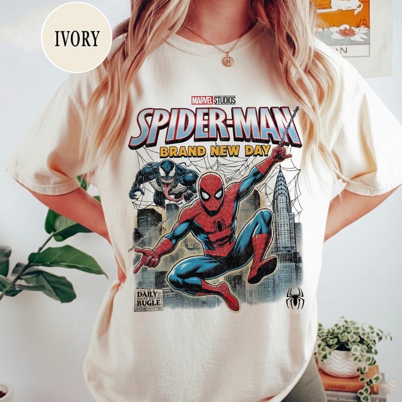 K&ouml;nnte beinhalten: Elfenbeinfarbenes T-Shirt mit einem Marvel Studios Spider-Man-Grafikdesign. Das Design zeigt Spider-Man, Venom, Stadtlandschaften und den Text "BRAND NEW DAY". Das Daily Bugle-Logo und ein Spinnensymbol sind ebenfalls zu sehen.