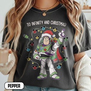 May include: Dark grey t-shirt with a Buzz Lightyear graphic wearing a Santa hat, encircled by Mickey Mouse Christmas lights. The text "TO INFINITY AND CHRISTMAS!" is above the graphic. The word "PEPPER" is at the bottom.