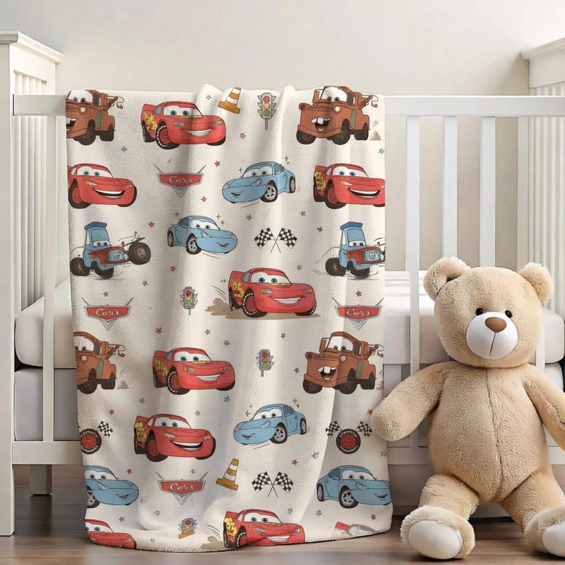 Puede incluir: Una manta de beb&eacute; color crema con un patr&oacute;n repetido de coches animados en rojo, azul y marr&oacute;n. La manta presenta im&aacute;genes de banderas de carreras, sem&aacute;foros y la palabra "Cars". Un oso de peluche est&aacute; al lado de la cuna.