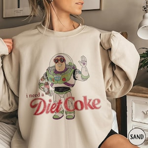 K&ouml;nnte beinhalten: Sandfarbenes Sweatshirt mit einem Cartoon-Bild von Buzz Lightyear, der eine Diet Coke-Flasche h&auml;lt. Der Text "i need a Diet Coke" ist in roter Schrift darunter gedruckt. Das Sweatshirt hat lange &Auml;rmel.
