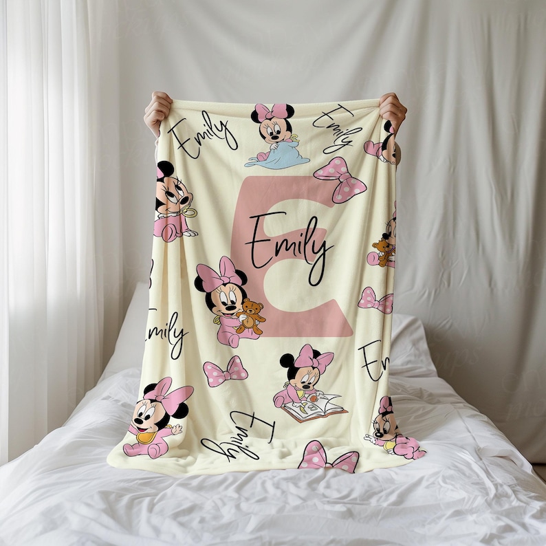 May include: A cream-colored baby blanket featuring a pink Minnie Mouse design with bows and the name "Emily" in a large pink letter and script. The blanket is decorated with multiple images of Minnie Mouse in various poses.