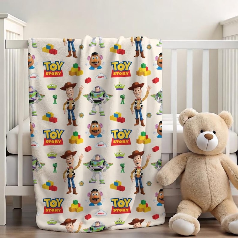May include: A cream-colored Toy Story blanket featuring Woody, Buzz Lightyear, Mr. Potato Head, and other characters. The blanket has a repeating pattern of the characters, blocks, and the Toy Story logo. A beige teddy bear sits next to the crib.