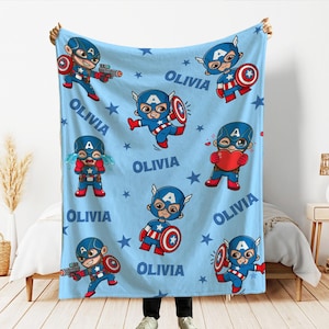 Pode incluir: Um cobertor azul claro com ilustra&ccedil;&otilde;es de desenhos animados do Capit&atilde;o Am&eacute;rica em v&aacute;rias poses, o nome "OLIVIA" e padr&otilde;es de estrelas. Os personagens do Capit&atilde;o Am&eacute;rica s&atilde;o em vermelho, branco e azul.