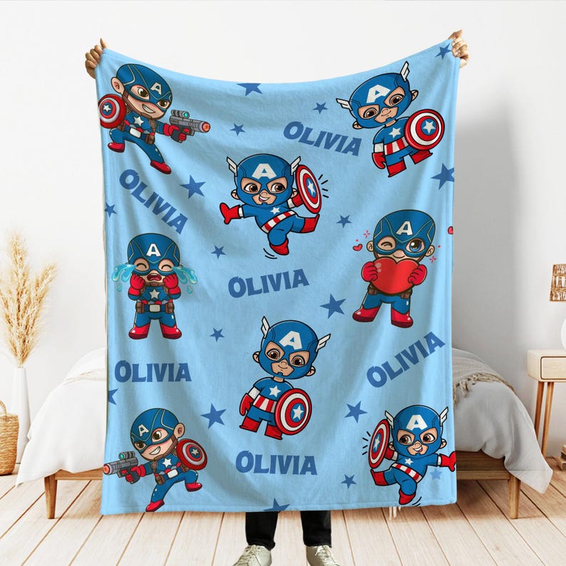 Pode incluir: Um cobertor azul claro com ilustra&ccedil;&otilde;es de desenhos animados do Capit&atilde;o Am&eacute;rica em v&aacute;rias poses, o nome "OLIVIA" e padr&otilde;es de estrelas. Os personagens do Capit&atilde;o Am&eacute;rica s&atilde;o em vermelho, branco e azul.