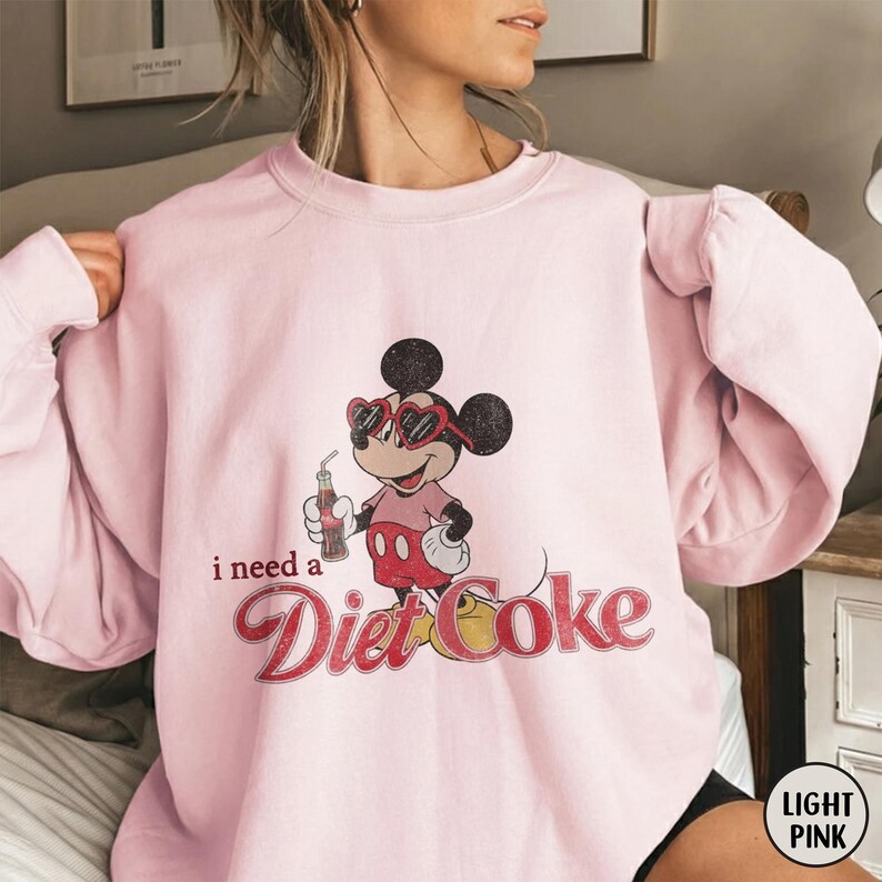 May include: Light pink sweatshirt featuring a vintage-style Mickey Mouse graphic wearing heart-shaped sunglasses and holding a Diet Coke. The text "i need a Diet Coke" is printed below the character in a retro font.