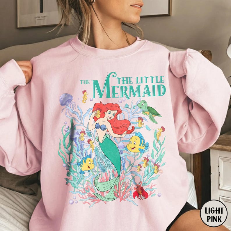 Puede incluir: Sudadera rosa claro con un dise&ntilde;o de Ariel de La Sirenita. El dise&ntilde;o incluye el texto "The Little Mermaid" en verde azulado, junto con varias criaturas marinas. La sudadera es de cuello redondo.