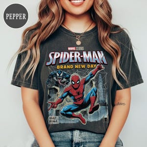 K&ouml;nnte beinhalten: Dunkelgraues T-Shirt mit einer Spider-Man-Grafik und dem Text "Marvel Studios Spider-Man Brand New Day". Das Design beinhaltet Venom und eine Stadtlandschaft. Das Daily Bugle-Logo ist ebenfalls sichtbar.