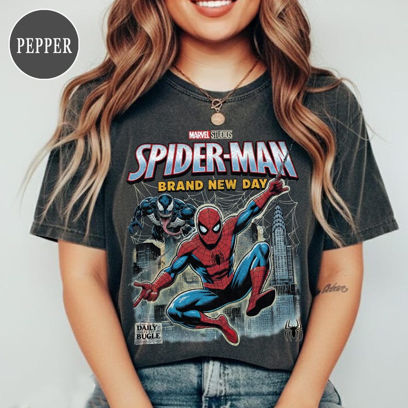 K&ouml;nnte beinhalten: Dunkelgraues T-Shirt mit einer Spider-Man-Grafik und dem Text "Marvel Studios Spider-Man Brand New Day". Das Design beinhaltet Venom und eine Stadtlandschaft. Das Daily Bugle-Logo ist ebenfalls sichtbar.