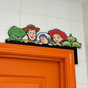 Puede incluir: Un adorno negro para la parte superior de la puerta con personajes de la franquicia de películas Toy Story. Los personajes incluyen a Rex el dinosaurio, Woody, Buzz Lightyear, Jessie y los extraterrestres de tres ojos. Montado en una puerta naranja.