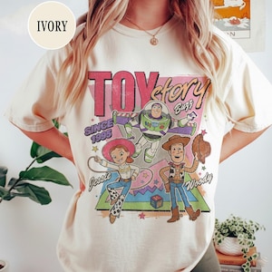 May include: Cream-coloured t-shirt with a Toy Story graphic featuring Woody, Buzz Lightyear, and Jessie. The design includes the text "TOY STORY" and "SINCE 1995". The shirt has a vintage, distressed appearance.