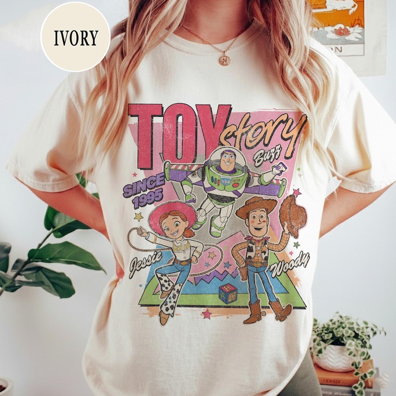 May include: Cream-coloured t-shirt with a Toy Story graphic featuring Woody, Buzz Lightyear, and Jessie. The design includes the text "TOY STORY" and "SINCE 1995". The shirt has a vintage, distressed appearance.