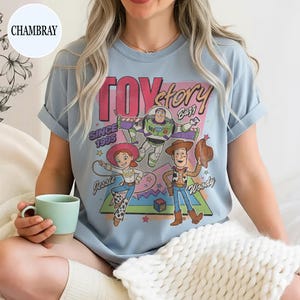 May include: Pale blue t-shirt with a graphic of Toy Story characters: Woody, Buzz Lightyear, and Jessie. The design includes the text "Toy Story" and "Since 1995". The sleeves are rolled up.