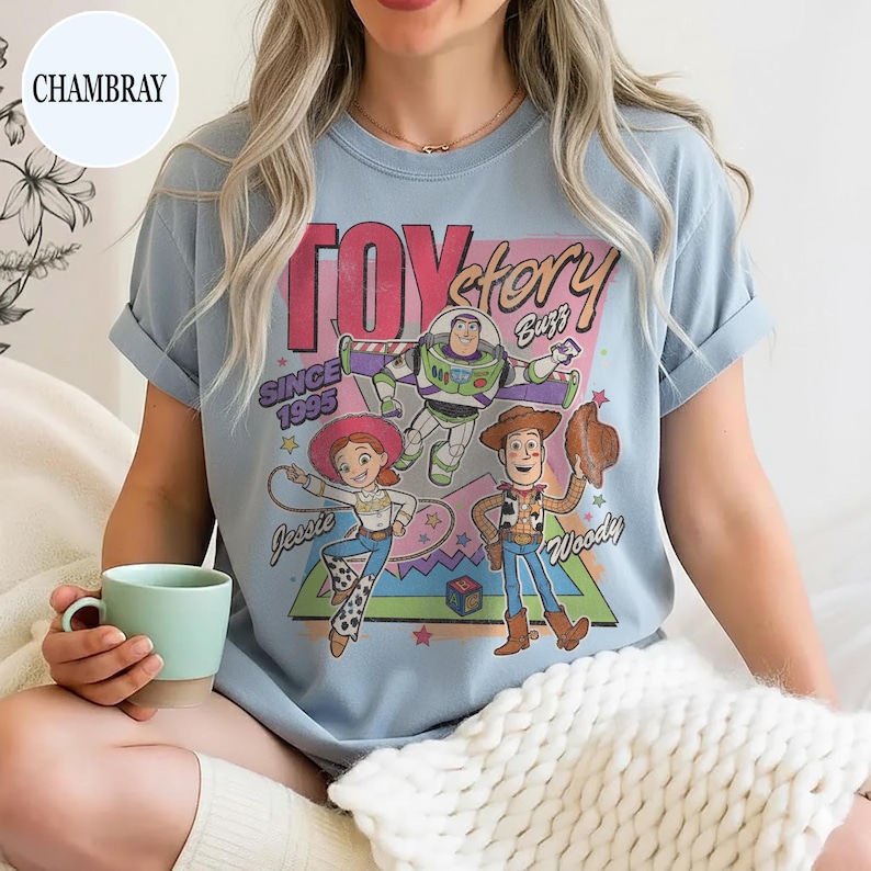 May include: Pale blue t-shirt with a graphic of Toy Story characters: Woody, Buzz Lightyear, and Jessie. The design includes the text "Toy Story" and "Since 1995". The sleeves are rolled up.