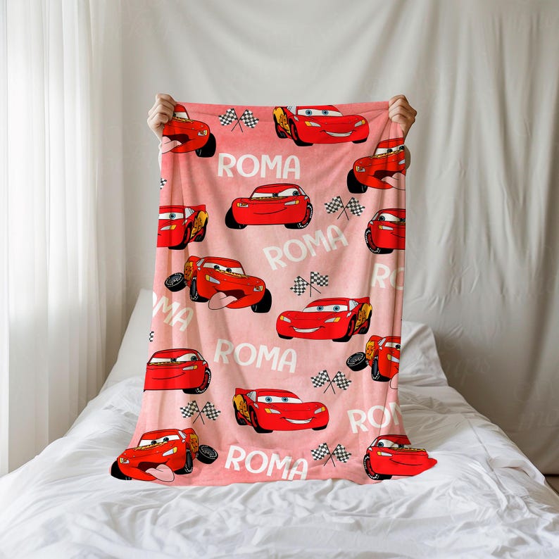 Puede incluir: Una manta rosa con un patr&oacute;n repetido de coches de carreras rojos de dibujos animados, banderas a cuadros y la palabra "ROMA" en blanco. La manta se muestra sobre un fondo blanco.