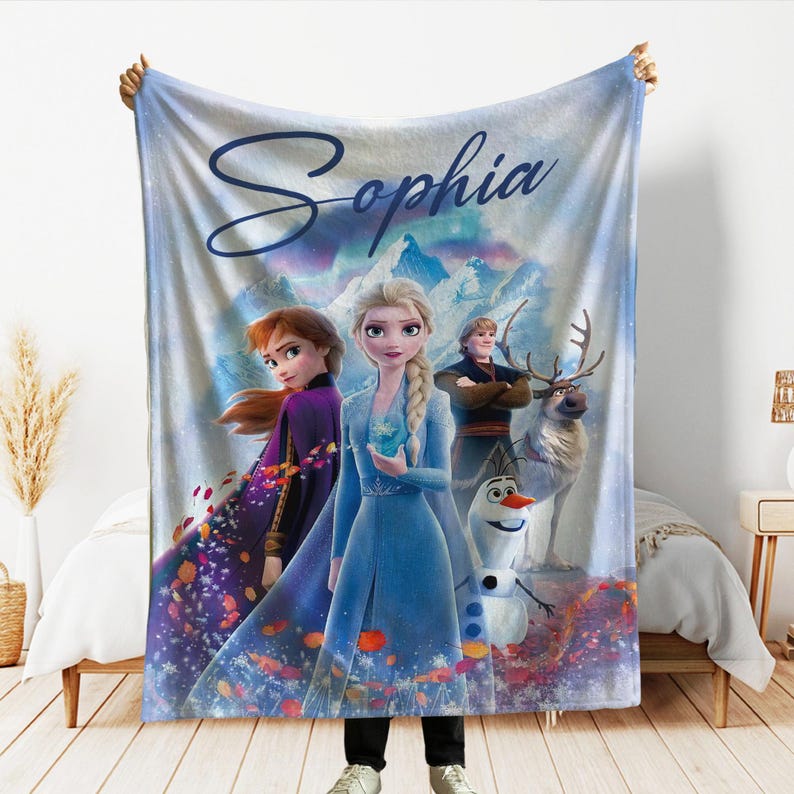K&ouml;nnte beinhalten: Eine personalisierte Fleece-Decke mit Figuren aus dem Film Frozen, darunter Elsa, Anna, Kristoff, Olaf und Sven. Die Decke ist hellblau mit einer Berglandschaft und dem Namen "Sophia".