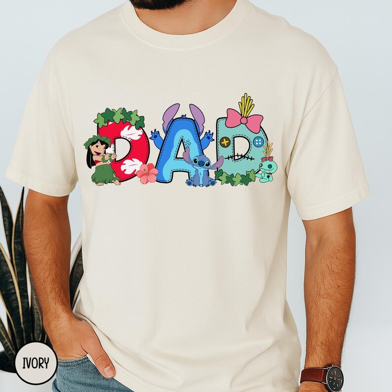 K&ouml;nnte beinhalten: Cremefarbenes T-Shirt mit dem Wort "DAD" in Comic-Schrift. Das "D" ist rot mit einem hawaiianischen M&auml;dchen, das "A" ist blau mit Stitch und das letzte "D" ist t&uuml;rkis. Die Buchstaben sind mit Figuren und floralen Elementen verziert.