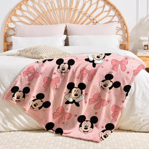 Op de afbeelding: Roze sprei met een herhaald patroon van Mickey Mouse-gezichten, roze strikken en kleine sterren. De sprei is gedrapeerd over een bed met wit beddengoed en een geweven hoofdbord.