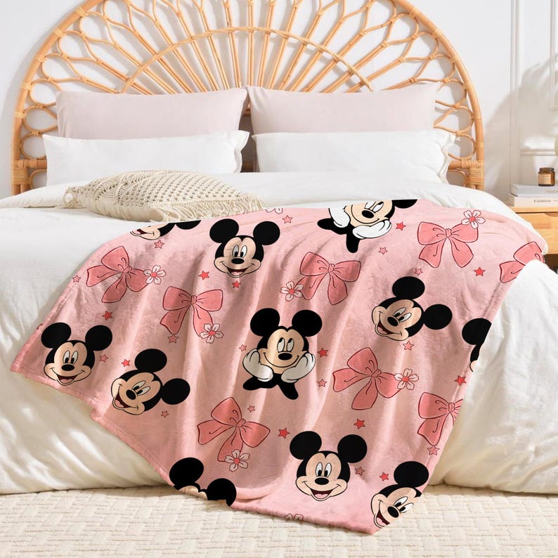 Op de afbeelding: Roze sprei met een herhaald patroon van Mickey Mouse-gezichten, roze strikken en kleine sterren. De sprei is gedrapeerd over een bed met wit beddengoed en een geweven hoofdbord.