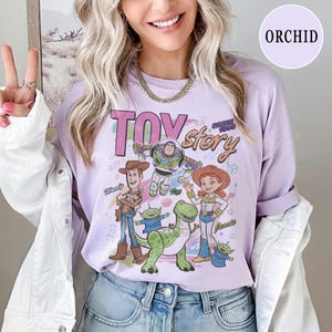 May include: A light purple long-sleeve shirt featuring the text "TOY STORY" and various cartoon characters. The shirt has a crew neck and a relaxed fit. The characters include Woody, Buzz Lightyear, Jessie, and others.