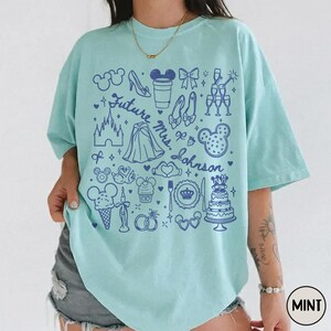 May include: A mint green short-sleeve t-shirt featuring a dark blue graphic print of various icons including a castle, wedding cake, rings, and the text Future Mrs. Johnson. The design includes outlines of ears, a glass slipper, and champagne flutes.