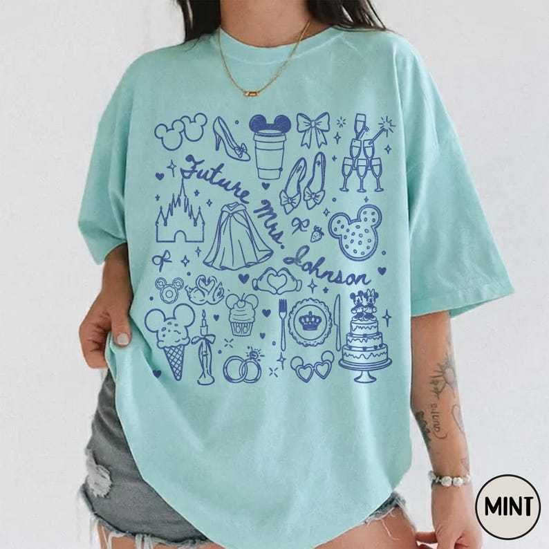 May include: A mint green short-sleeve t-shirt featuring a dark blue graphic print of various icons including a castle, wedding cake, rings, and the text Future Mrs. Johnson. The design includes outlines of ears, a glass slipper, and champagne flutes.