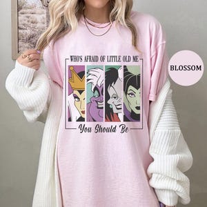 Puede incluir: Camiseta rosa claro con un gr&aacute;fico de cuatro villanos de dibujos animados y el texto "WHO'S AFRAID OF LITTLE OLD ME - YOU SHOULD BE". La camiseta tiene cuello redondo y mangas cortas.