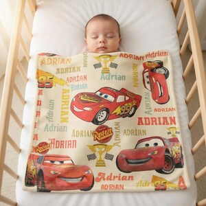 Op de afbeelding: Een beige babydeken met een rood cartoon raceauto-ontwerp en de naam "Adrian" erop gedrukt. De deken heeft rode, gele en zwarte accenten. De baby slaapt in een houten bedje.