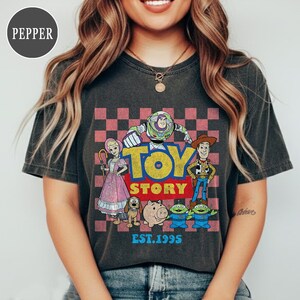 以下が含まれることがあります： トイ・ストーリーのロゴと、ウッディやバズ・ライトイヤーなどのキャラクターが描かれた濃いグレーのTシャツ。ピンクと赤のチェック柄の背景。ロゴの下には「EST.1995」の文字。