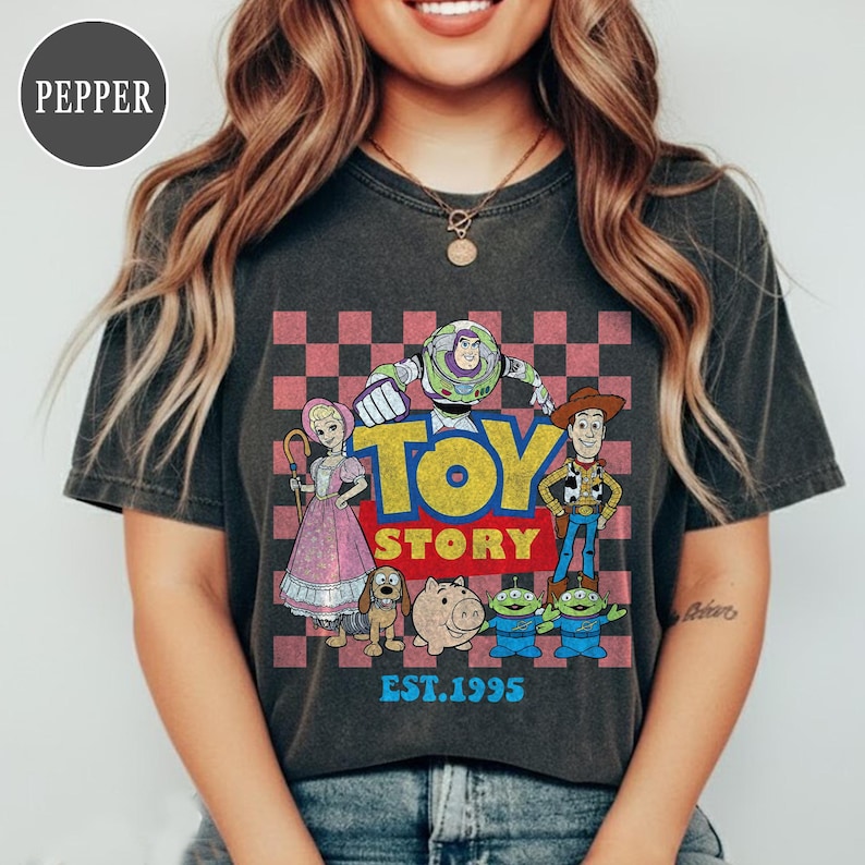 以下が含まれることがあります： トイ・ストーリーのロゴと、ウッディやバズ・ライトイヤーなどのキャラクターが描かれた濃いグレーのTシャツ。ピンクと赤のチェック柄の背景。ロゴの下には「EST.1995」の文字。