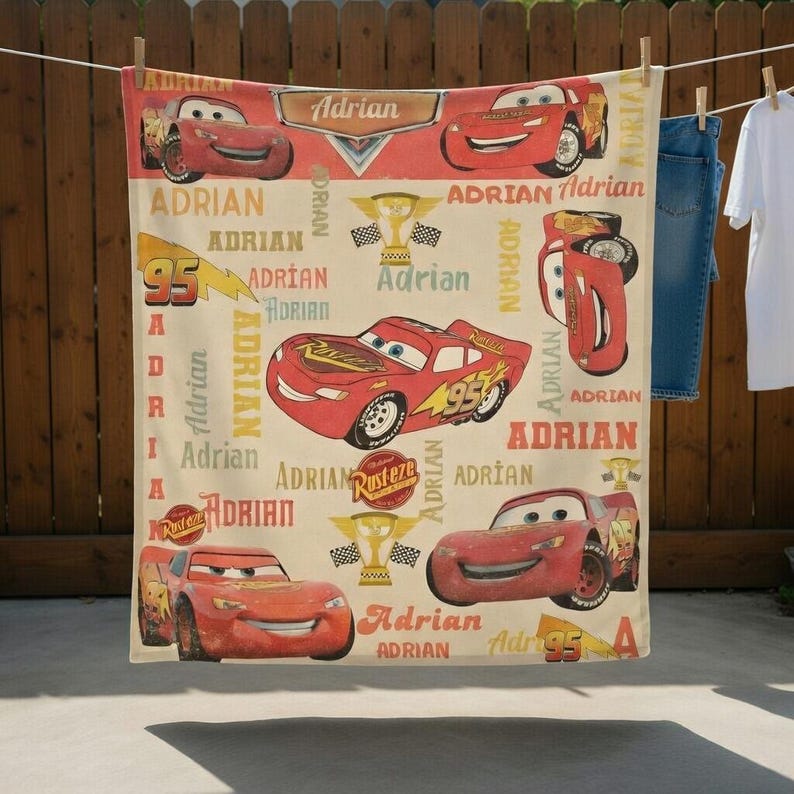 Op de afbeelding: Een beige deken met een herhalend patroon van het Lightning McQueen-personage uit de film Cars, het nummer 95 en de naam "Adrian" in het rood. De deken hangt aan een waslijn, met andere kleding op de achtergrond.