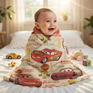 Op de afbeelding: Een baby gewikkeld in een beige deken met rode cartoon raceauto's en de naam "Adrian" in verschillende lettertypen. De deken heeft een zachte textuur en is versierd met race-thema graphics. Speelgoed is zichtbaar op de achtergrond.