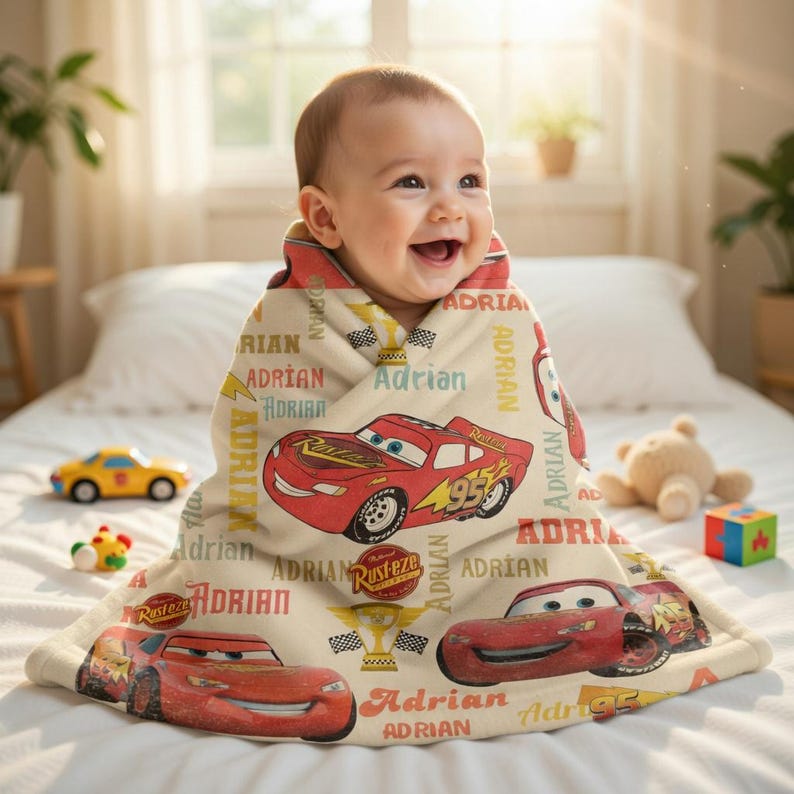 Op de afbeelding: Een baby gewikkeld in een beige deken met rode cartoon raceauto's en de naam "Adrian" in verschillende lettertypen. De deken heeft een zachte textuur en is versierd met race-thema graphics. Speelgoed is zichtbaar op de achtergrond.