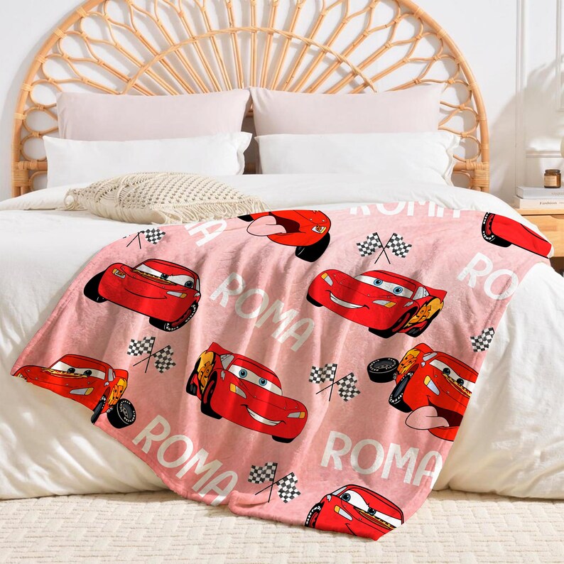 Puede incluir: Una manta de forro polar rosa con un patr&oacute;n repetido de coches de carreras rojos, banderas a cuadros y la palabra "ROMA" en blanco. La manta est&aacute; sobre una cama con un cabecero tejido.