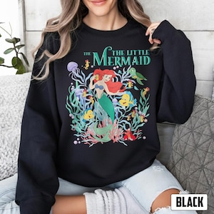 Puede incluir: Sudadera negra con un gr&aacute;fico colorido de Ariel de "La Sirenita". El dise&ntilde;o incluye a Ariel con cabello rojo y una cola verde, rodeada de criaturas marinas y el texto "The Little Mermaid."