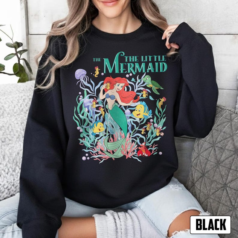 Puede incluir: Sudadera negra con un gr&aacute;fico colorido de Ariel de "La Sirenita". El dise&ntilde;o incluye a Ariel con cabello rojo y una cola verde, rodeada de criaturas marinas y el texto "The Little Mermaid."
