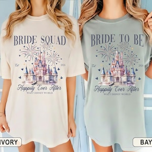 Puede incluir: Camisetas de color marfil y bahía con el texto "BRIDE SQUAD" y "BRIDE TO BE" en una fuente estilizada. Cada camiseta presenta una ilustración de acuarela de un castillo, fuegos artificiales y las palabras "Happily Ever After" y "Walt Disney World".