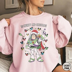 May include: Pale pink sweatshirt with a Buzz Lightyear graphic wearing a Santa hat and the text "TO INFINITY AND CHRISTMAS!". The design includes a string of lights with Mickey Mouse heads.