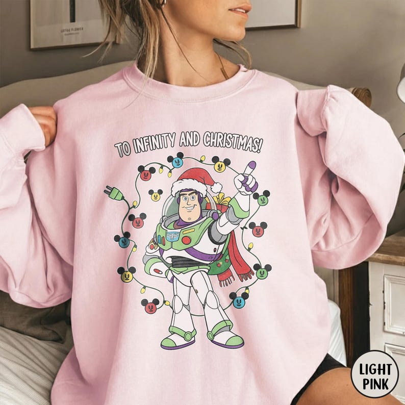 May include: Pale pink sweatshirt with a Buzz Lightyear graphic wearing a Santa hat and the text "TO INFINITY AND CHRISTMAS!". The design includes a string of lights with Mickey Mouse heads.