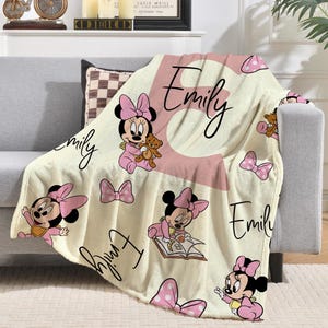 May include: Cream-colored baby blanket featuring Minnie Mouse illustrations in pink. The blanket has the name "Emily" in a cursive font and a large pink letter "E". Pink bows and polka dots are also featured.