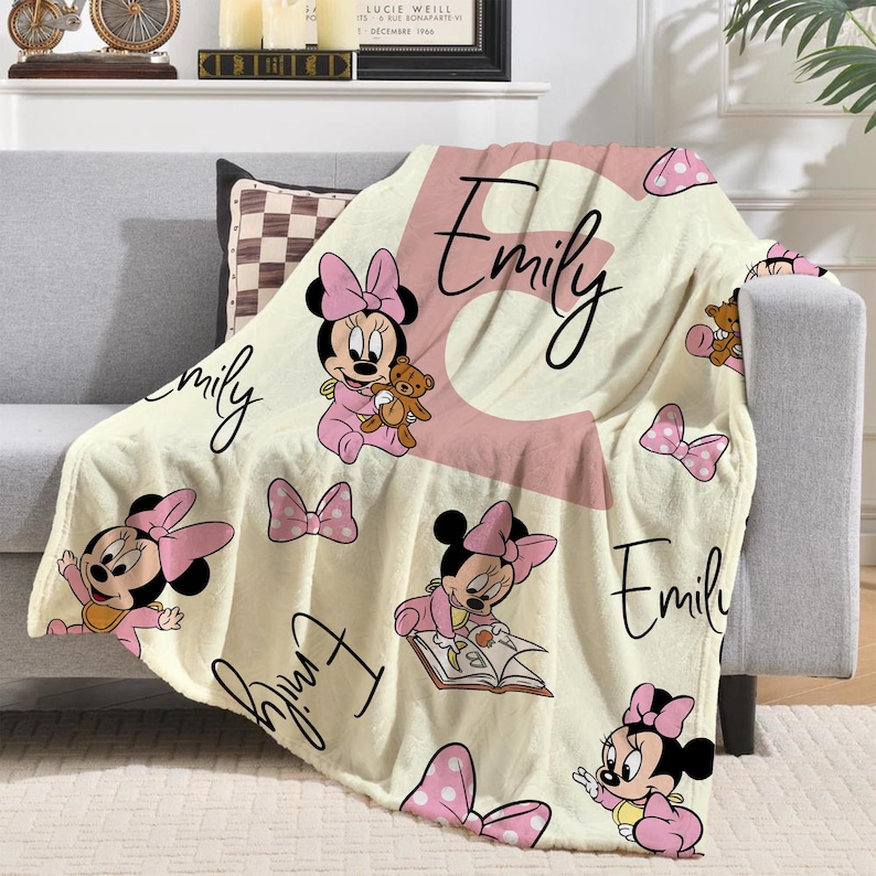 May include: Cream-colored baby blanket featuring Minnie Mouse illustrations in pink. The blanket has the name "Emily" in a cursive font and a large pink letter "E". Pink bows and polka dots are also featured.