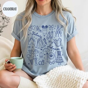 May include: A chambray blue short-sleeve t-shirt featuring a dark blue graphic print of various icons including a castle, wedding cake, rings, and the text "Future Mrs. Johnson" surrounded by small stars, hearts, and assorted wedding-themed symbols.