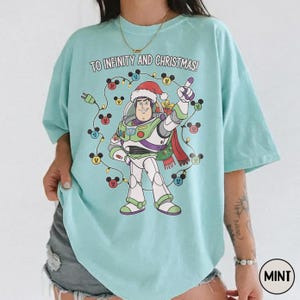 May include: A pale blue t-shirt with a Buzz Lightyear graphic wearing a Santa hat, encircled by Mickey Mouse-shaped Christmas lights. The text "TO INFINITY AND CHRISTMAS!" is printed above the character.