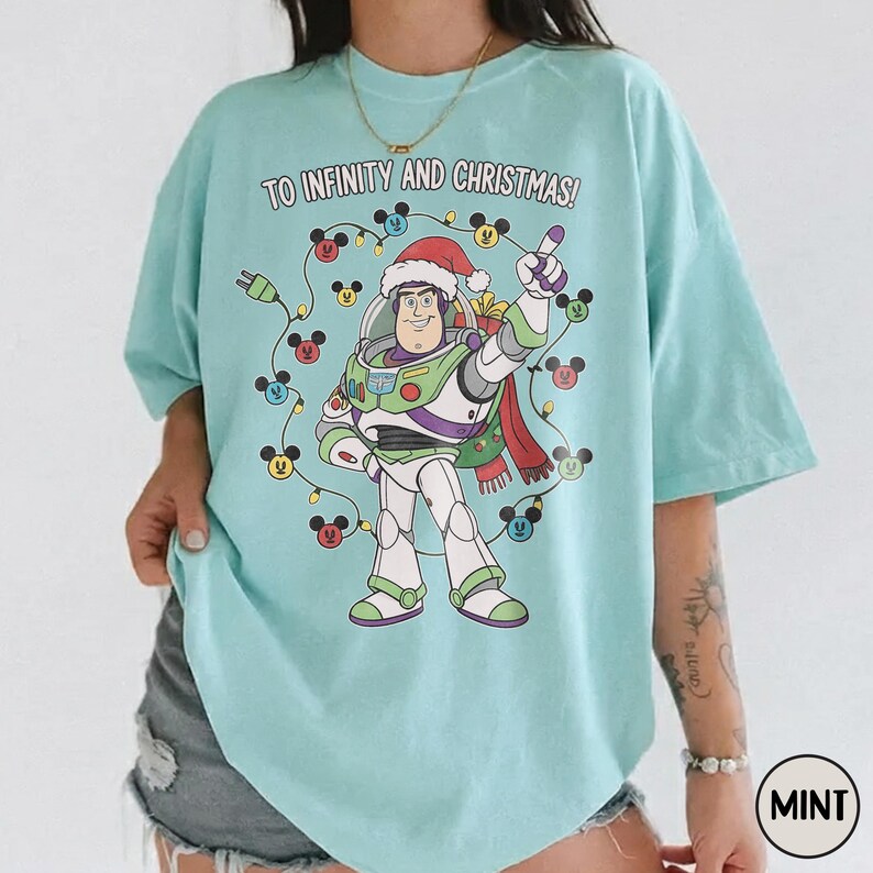May include: A pale blue t-shirt with a Buzz Lightyear graphic wearing a Santa hat, encircled by Mickey Mouse-shaped Christmas lights. The text "TO INFINITY AND CHRISTMAS!" is printed above the character.