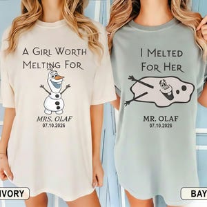 以下が含まれることがあります： 2枚の半袖Tシャツ。1枚はアイボリー、もう1枚はベイで、アナと雪の女王のオラフが描かれています。アイボリーのシャツには「A GIRL WORTH MELTING FOR」とオラフが描かれています。ベイのシャツには「I MELTED FOR HER」と溶けたオラフが描かれています。どちらのシャツにも「MRS. OLAF 07.10.2026」と「MR. OLAF 07.10.2026」と書かれています。