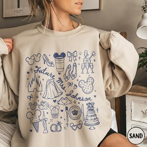 May include: A sand-coloured crewneck sweatshirt featuring a blue graphic print of various icons including a castle, glass slippers, a tiered cake, and the text Future Mrs. Johnson. The design includes small hearts, stars, and wedding-themed motifs.