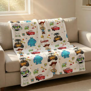 May include: White fleece blanket featuring cartoon characters from the movie Toy Story, including Woody, Buzz Lightyear, and Sully. The blanket also has the name "Lucas" printed on it. The blanket is draped over a beige sofa.